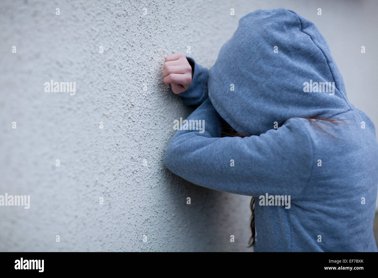 Sad child in hoodie with face hidden, leaning against a wall in despair Stock Photo