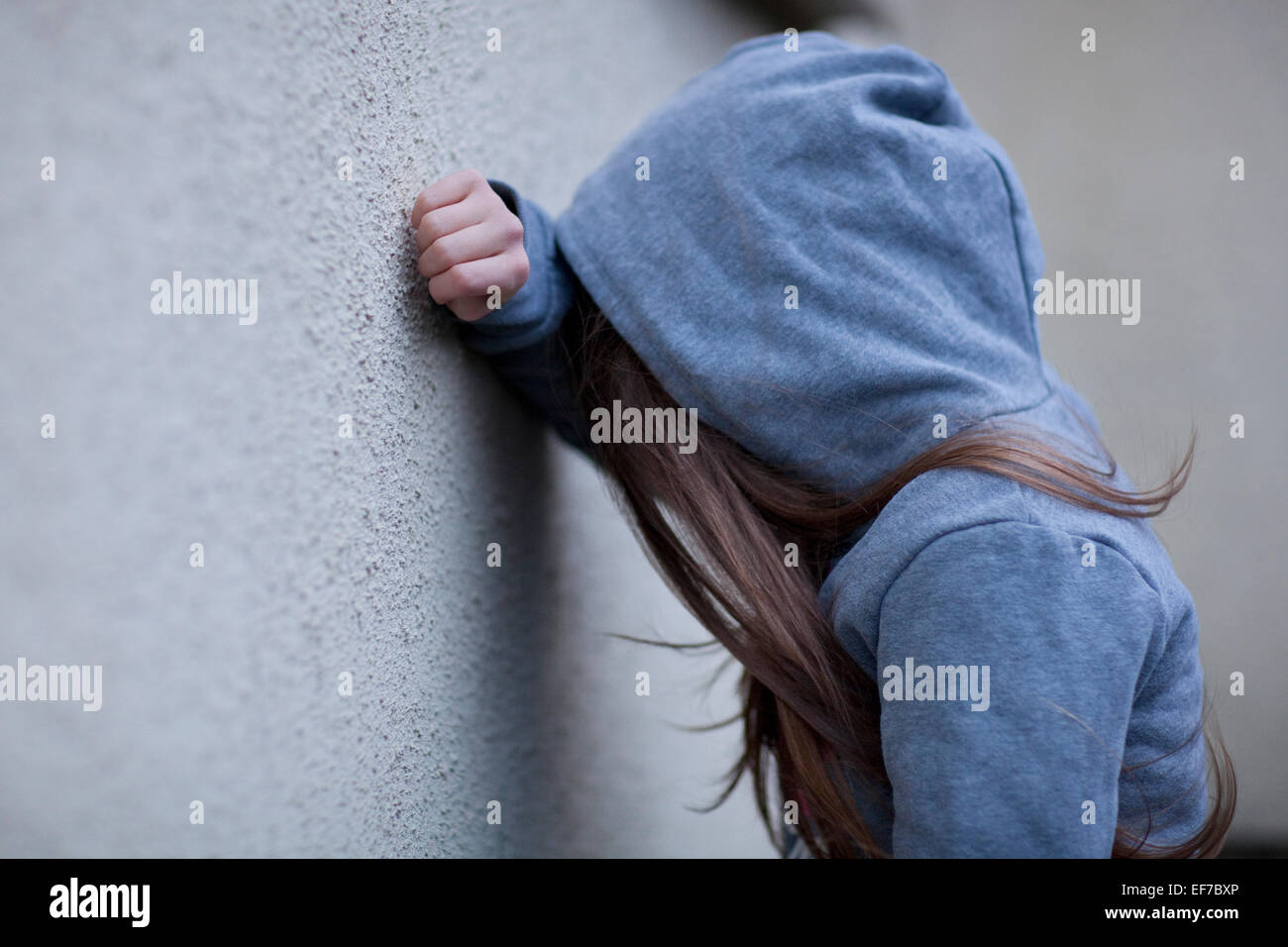 Sad girl in hoodie with face hidden, leaning against a wall in despair Stock Photo