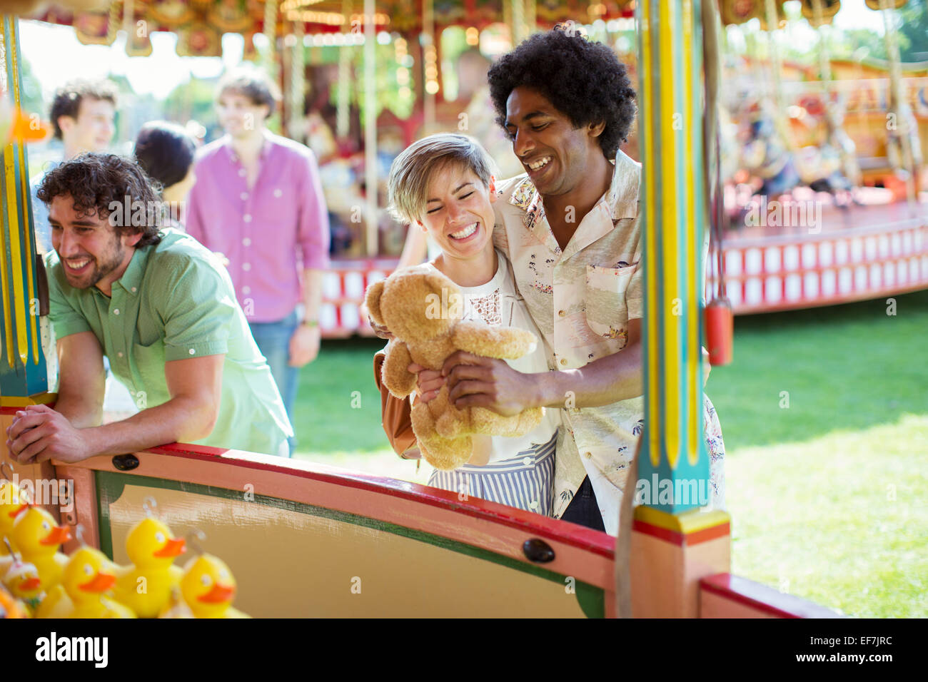 Smiling couple holding teddy bear next to fishing game in amusement park Stock Photo