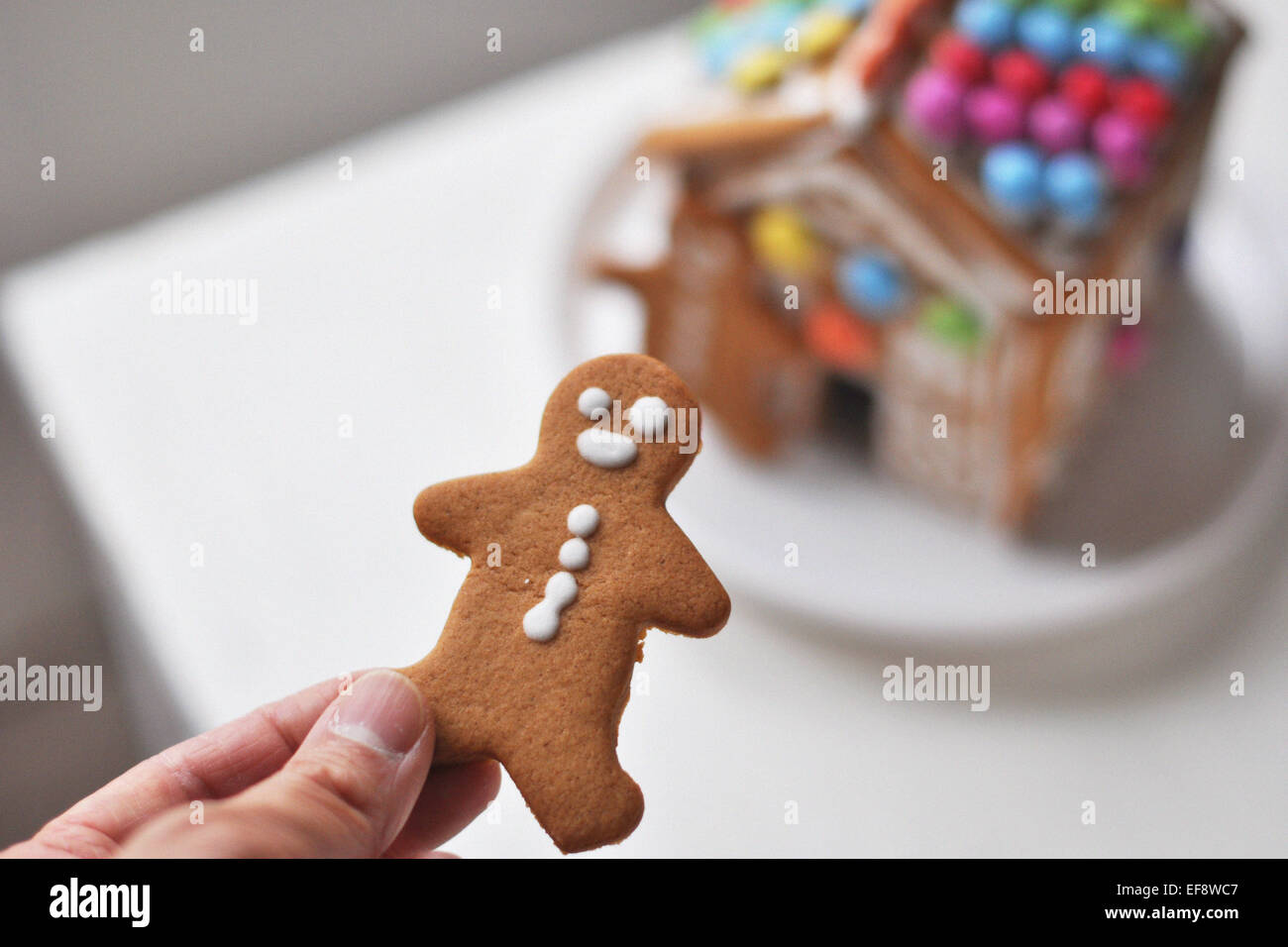Person's hand holding gingerbread man, gingerbread house in background Stock Photo