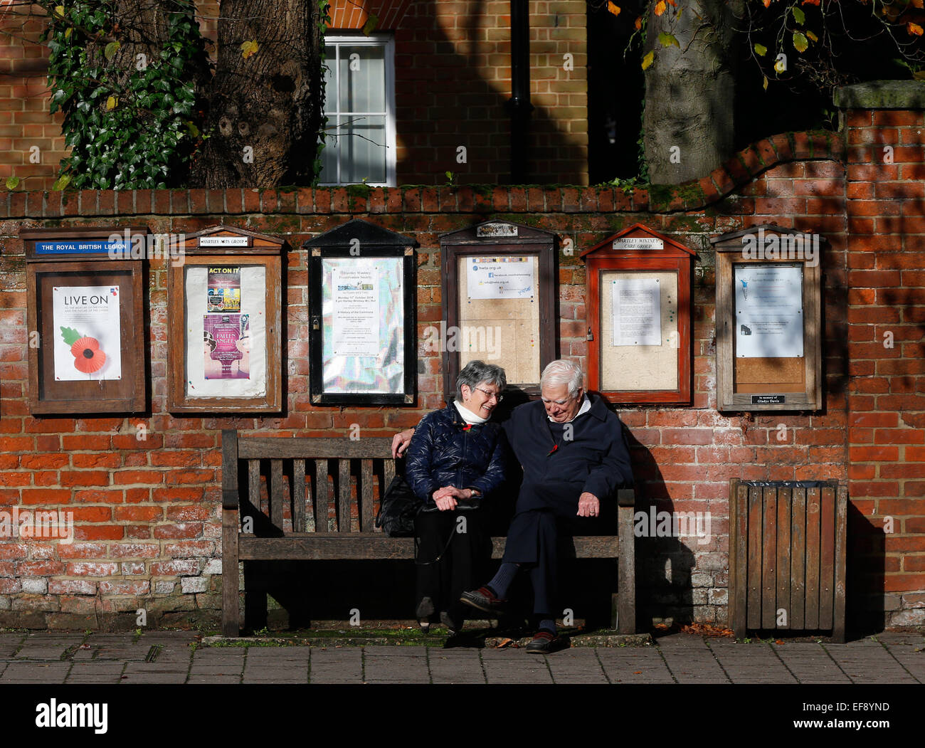 An elderly couple sit on a bench to have a conversation beneath notice boards in Hartley Wintney, Hook November 2014 Stock Photo