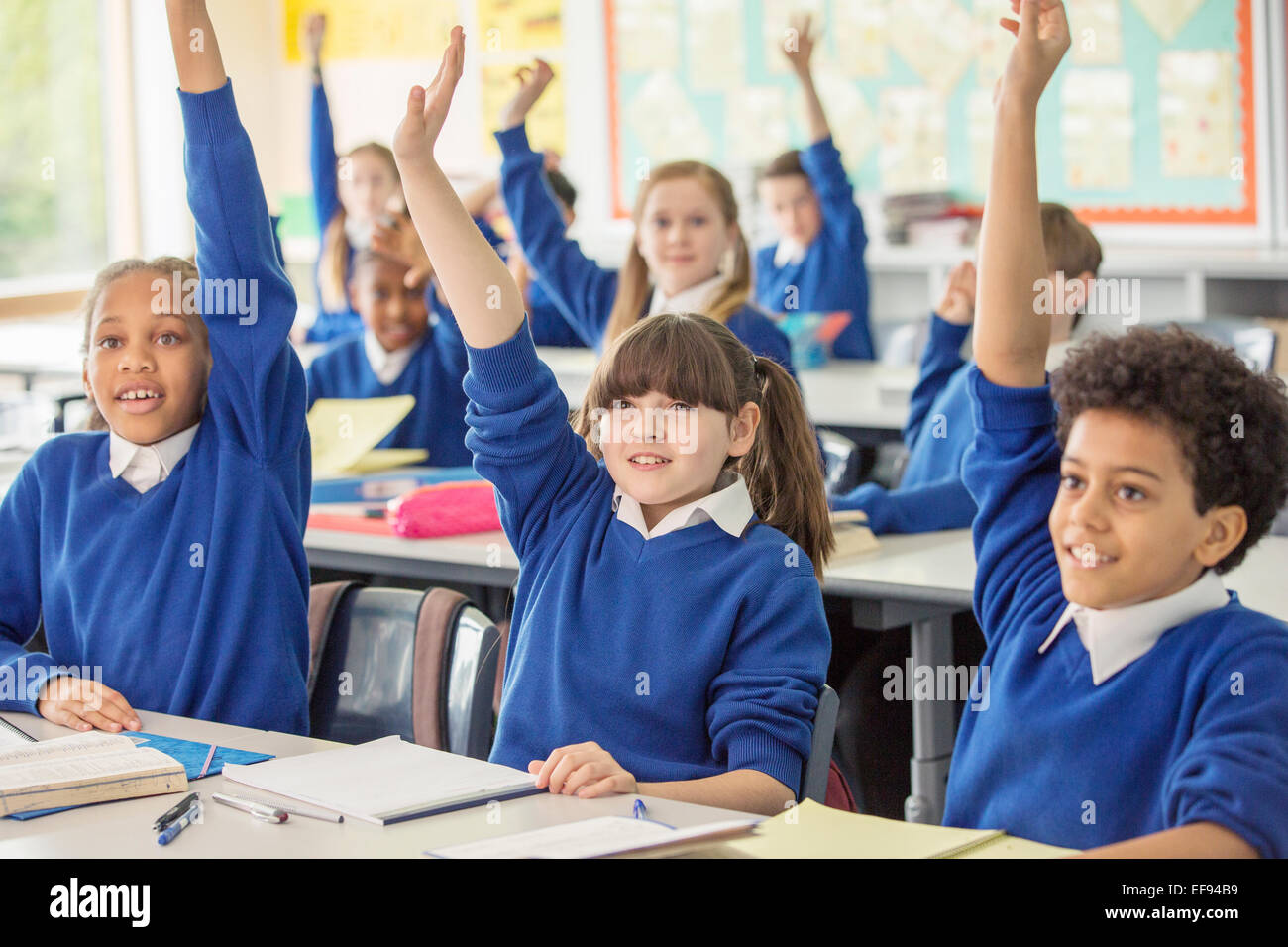 Elementary school children wearing blue school uniforms raising hands in classroom Stock Photo
