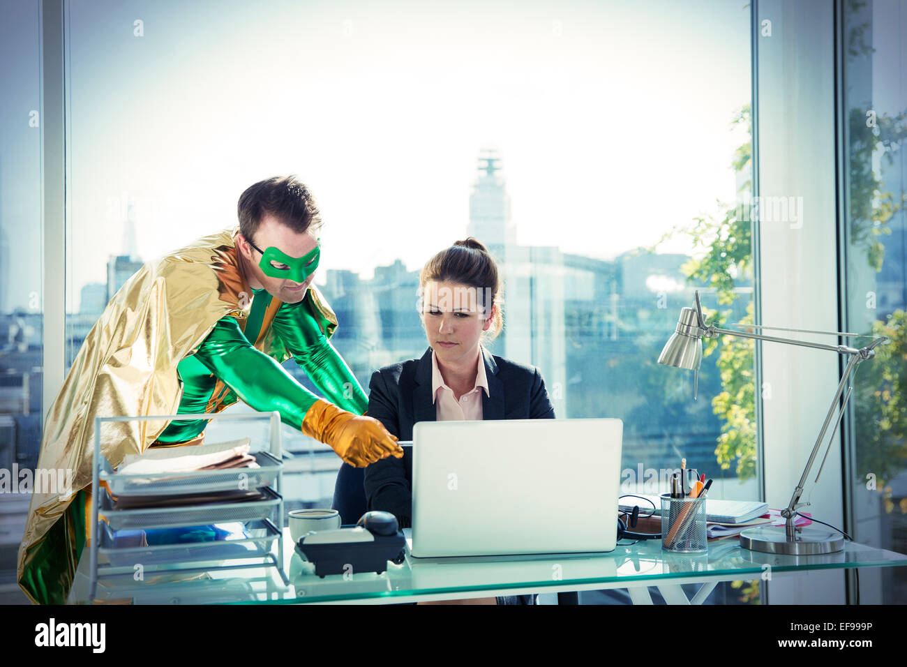 Superhero helping businesswoman working at office desk Stock Photo