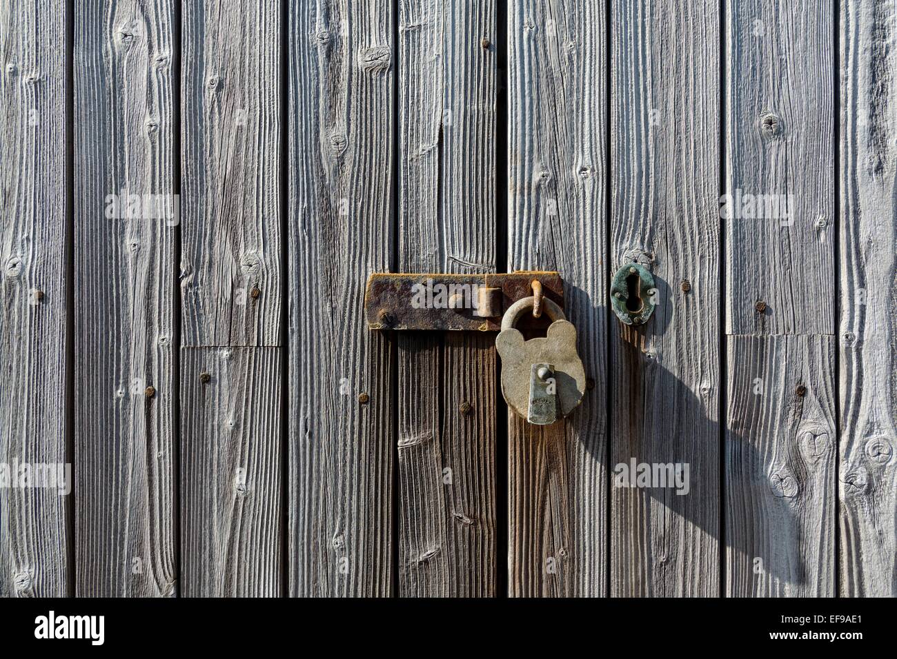 Old weathered and rusting metal latch and padlock on an old weathered wooded slatted door.  Picture includes a metal keyhole. Stock Photo