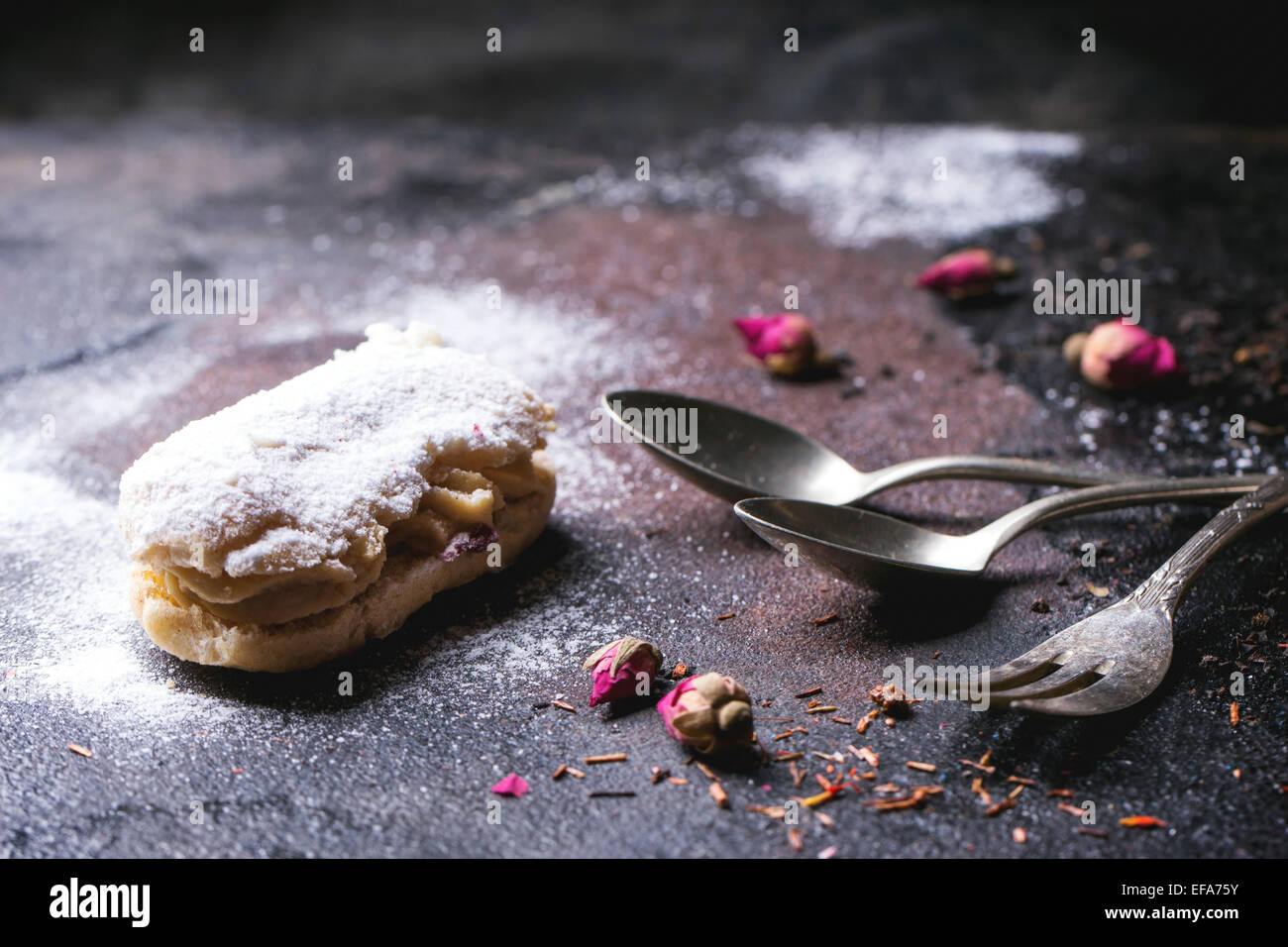 Eclairs with sugar powder, served with dry tea rose buds and vintage cutlery over dark background Stock Photo