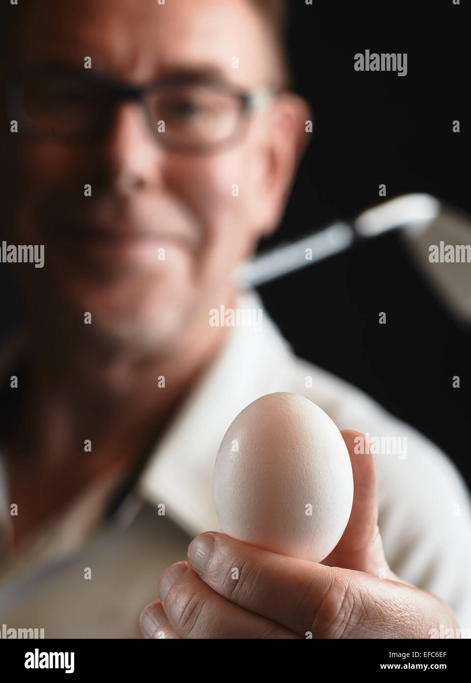 Close up, mature man golfer wearing a white shirt and he holds a white egg on hand, iron golf club on his shoulder - focus on ba Stock Photo
