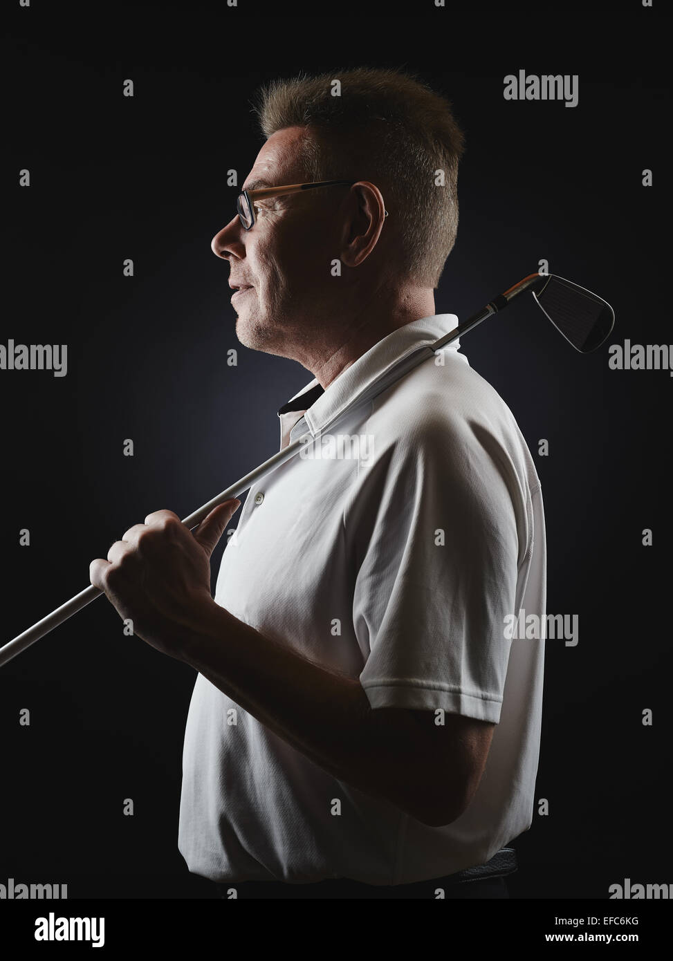 Side view, mature man golfer wearing a white shirt and he holds a iron golf club on his shoulder - studio shot, black background Stock Photo
