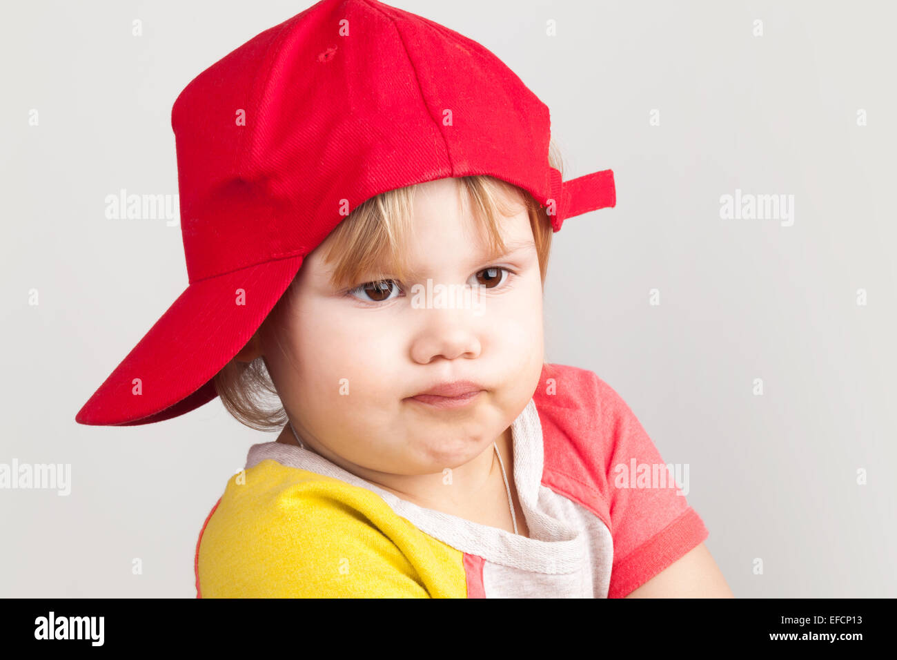 Studio portrait of funny confused baby girl in red baseball cap over gray wall background Stock Photo