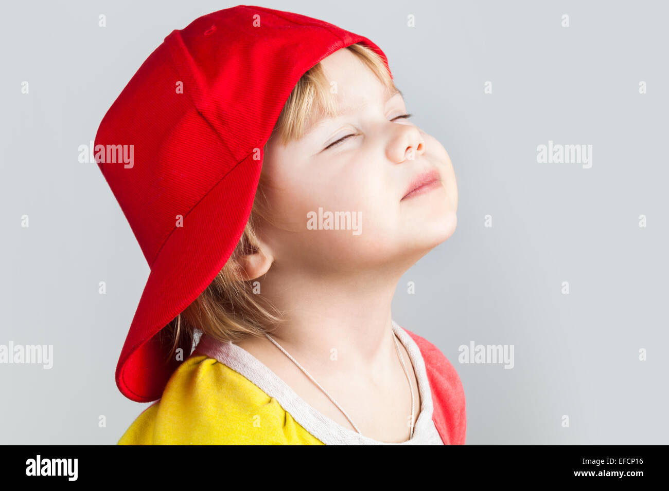 Studio portrait of happy baby girl with closed eyes in red baseball cap over gray wall background Stock Photo
