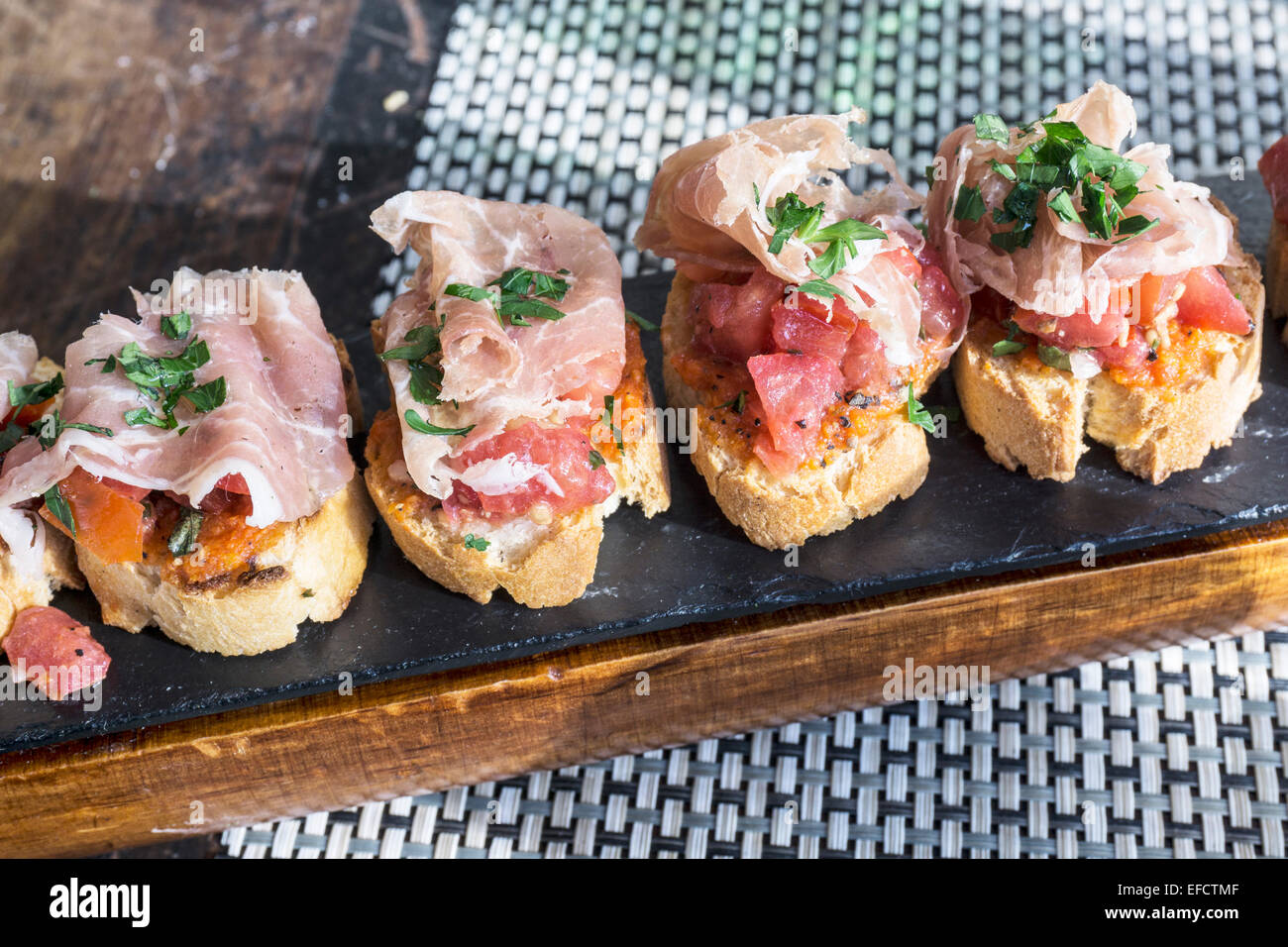 Bruschetta Serrano with Serrano ham fresh tomato & herbs on rounds of toasted baguette served on wooden board at outdoor table Stock Photo