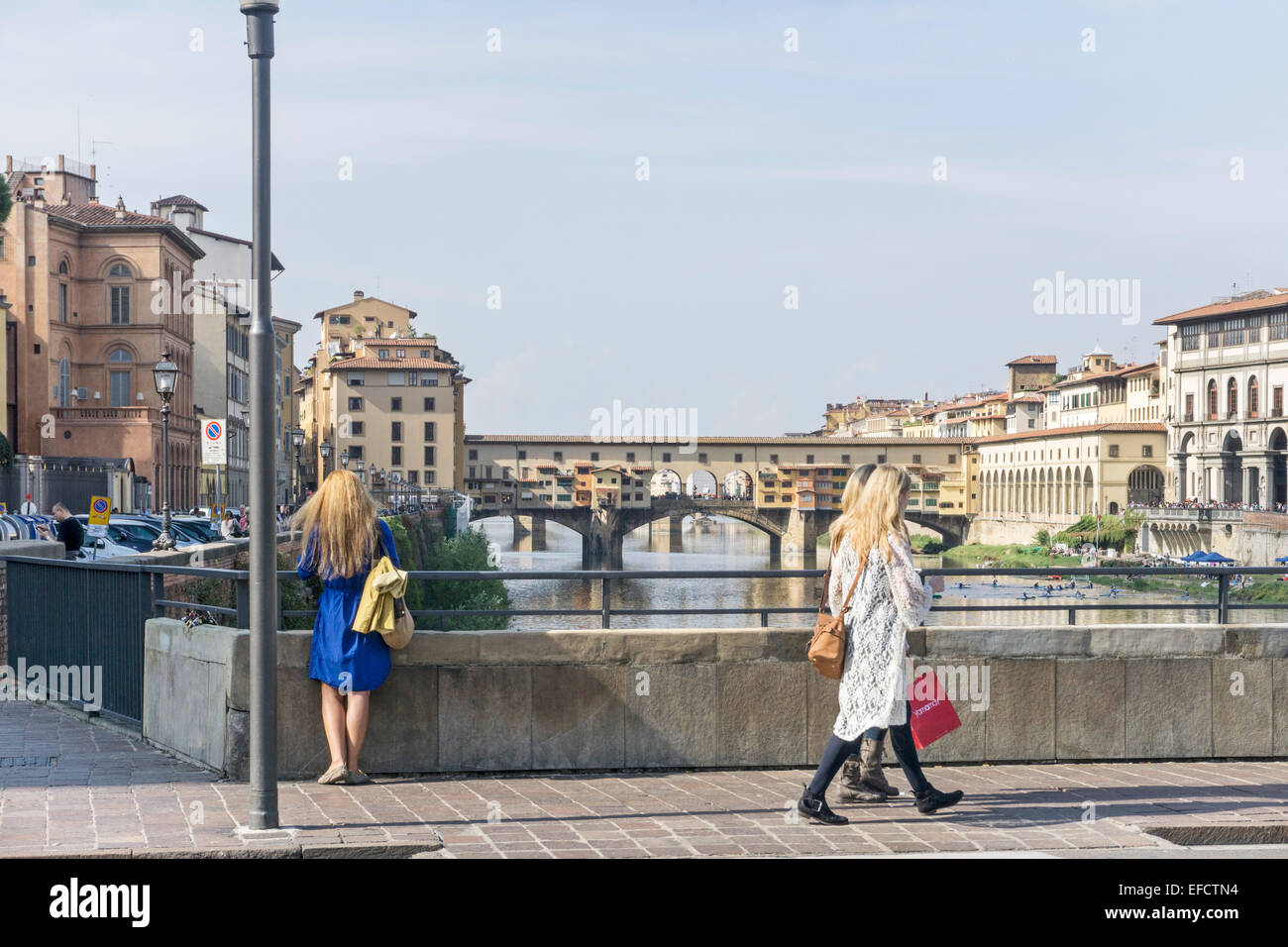 girl in blue dress with long fair hair stands at rail of Ponte alle Grazie gazing at Arno as 2 other pretty girls pass by Stock Photo