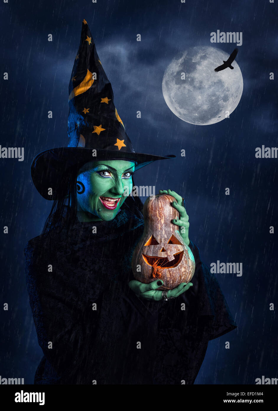 Witch with green skin holding carved Halloween pumpkin at moon and dark sky with rain Stock Photo