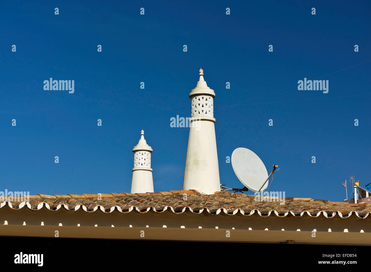 Two white chimneys against a deep blue sky in Olhos de Agua, Portugal. The terracotta tiled roof and a satellite dish are seen. Stock Photo
