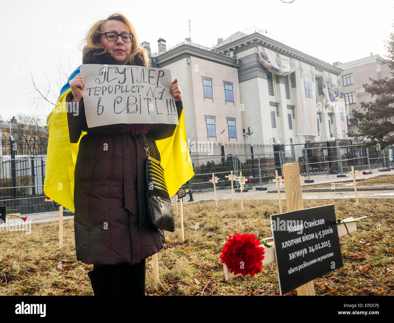 Crosses placed by activists and bearing the names of 30 people who died in shelling in Mariupol on Jan, 24 are seen in the foreground as woman standing with a banner 'Putler - №1 terrorist in the world' near the Russian Embassy in Kiev, Ukraine, Sunday, Feb. 1, 2015. Stock Photo