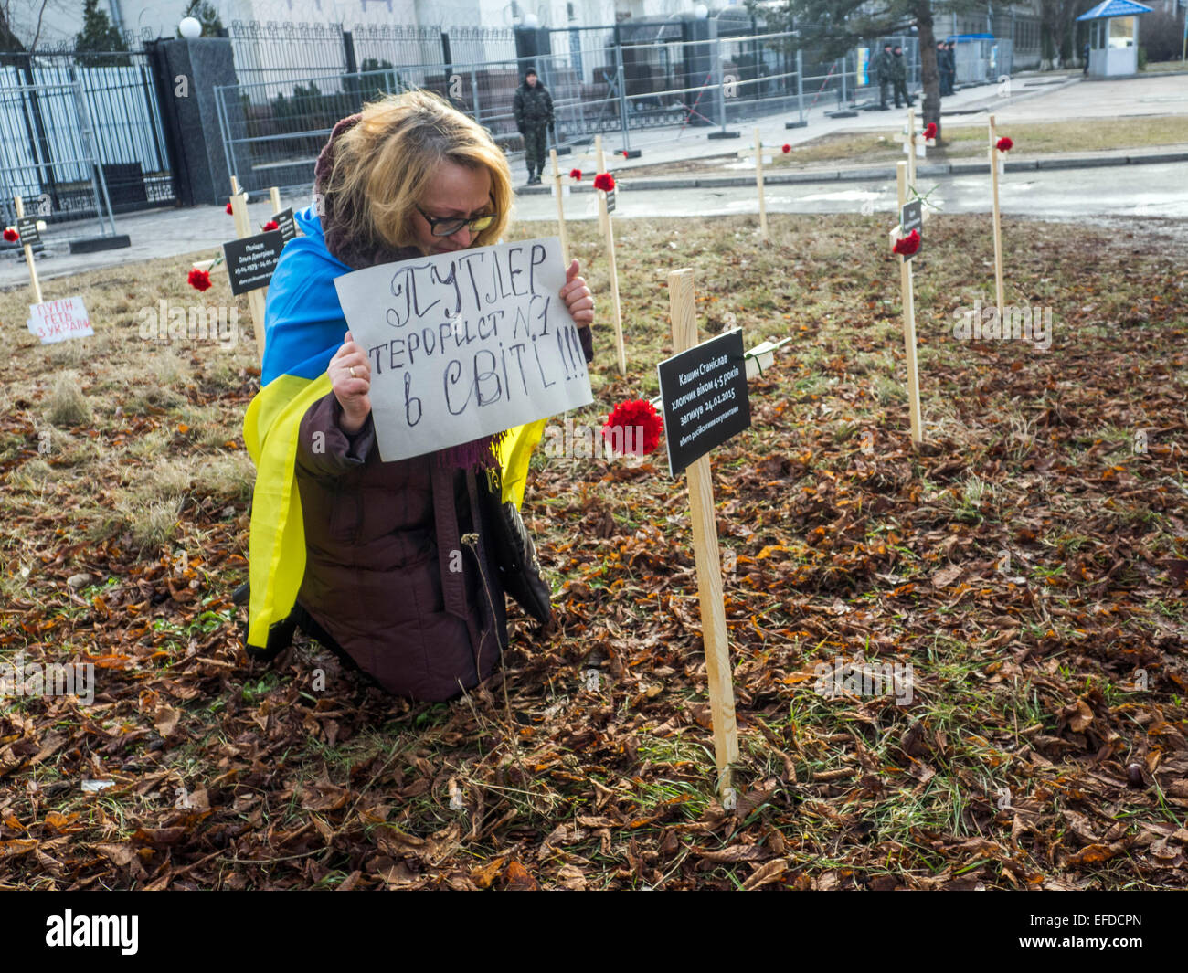 Crosses placed by activists and bearing the names of 30 people who died in shelling in Mariupol on Jan, 24 are seen in the foreground as woman kneeling with a banner 'Putler - №1 terrorist in the world' at the cross with the name of 4-5 year-old boy Stanislav Kashin, who was killed in Mariupol near the Russian Embassy in Kiev, Ukraine, Sunday, Feb. 1, 2015. Stock Photo