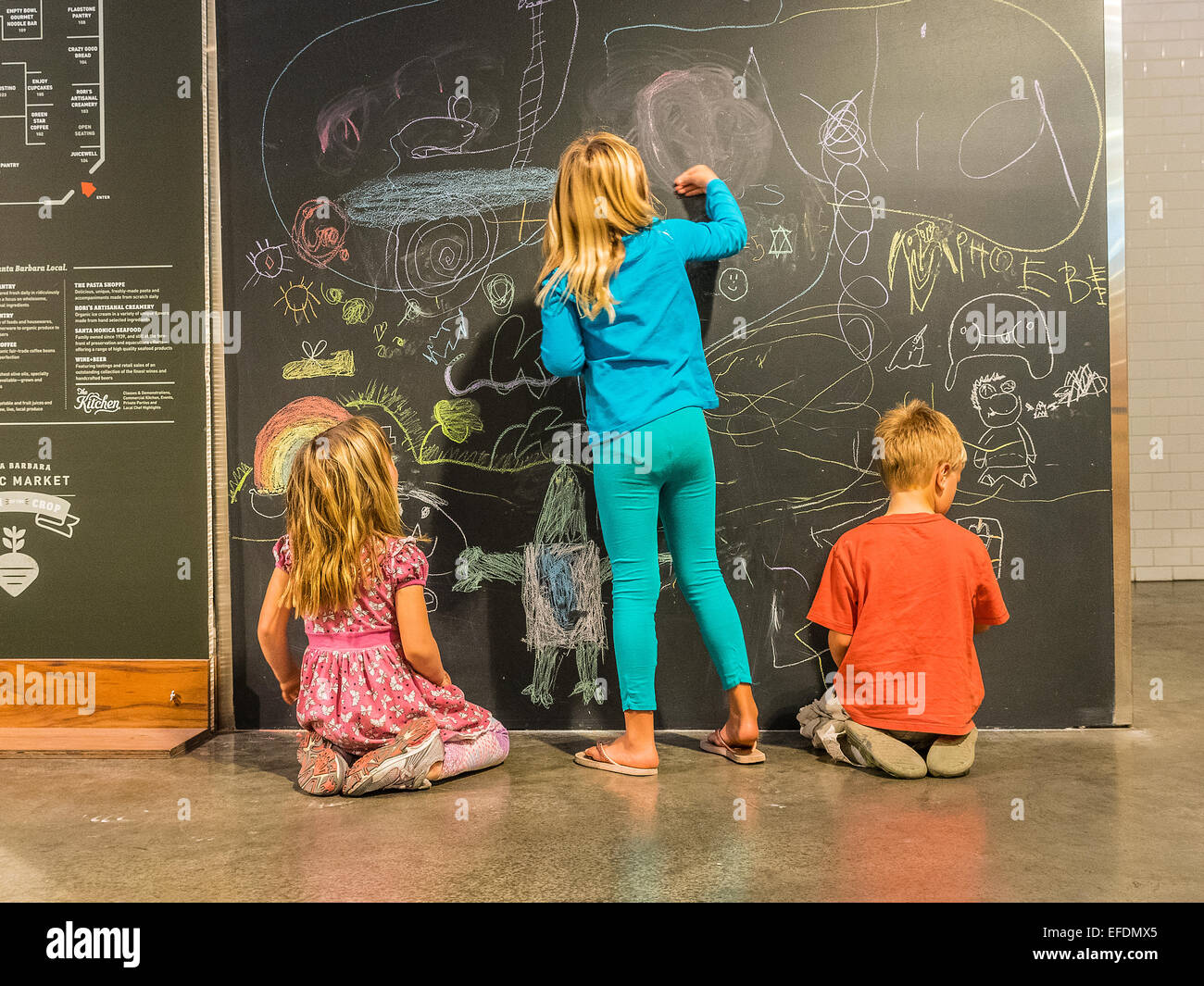 Three young siblings ages 5-10 years old draw with colored chalk on a blackboard in the Santa Barbara Public Market. Stock Photo