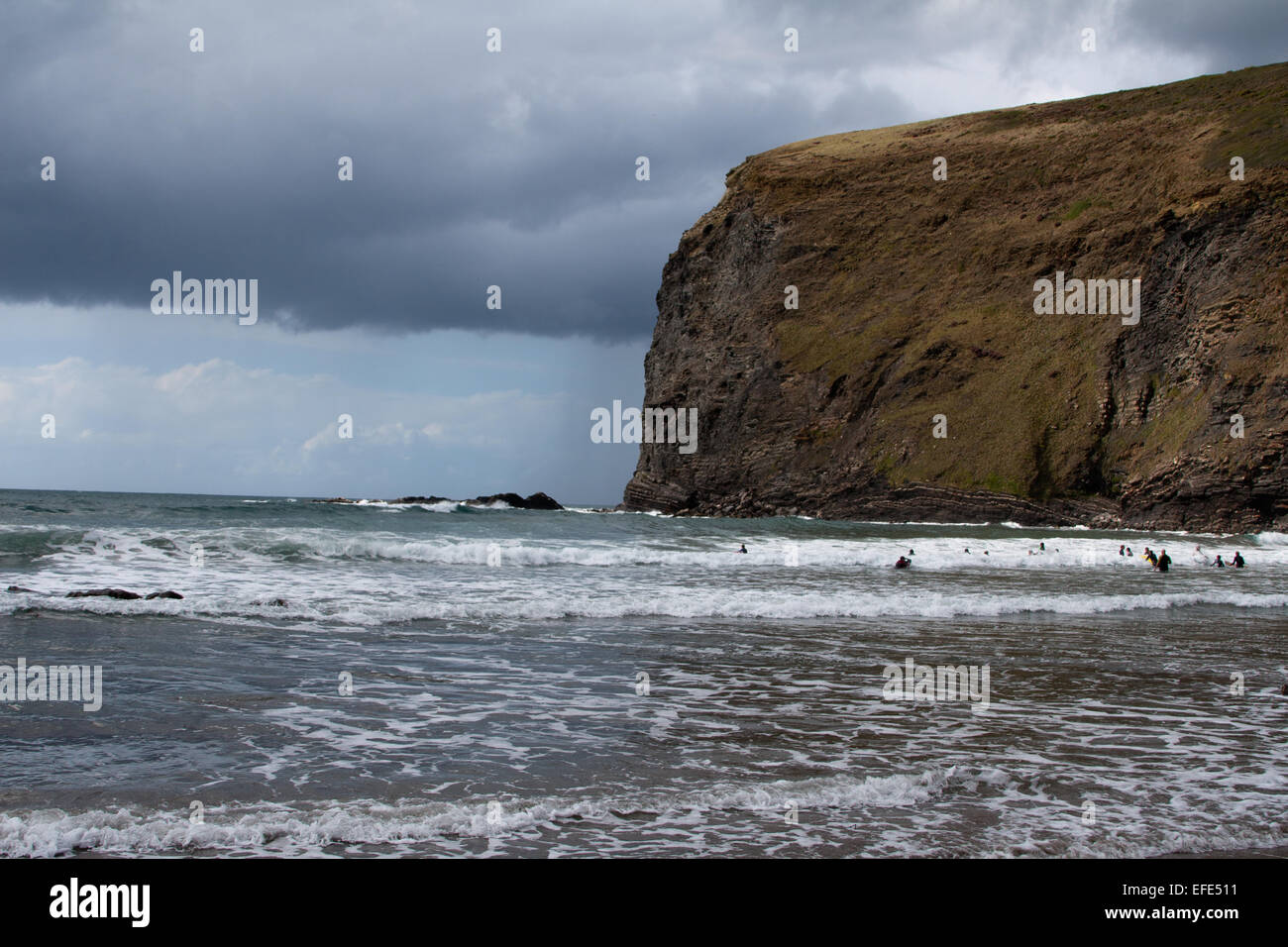 Swimmers in the sea near a rugged cliff under a cloudy sky on an overcast day at Crackington Haven, Cornwall Stock Photo