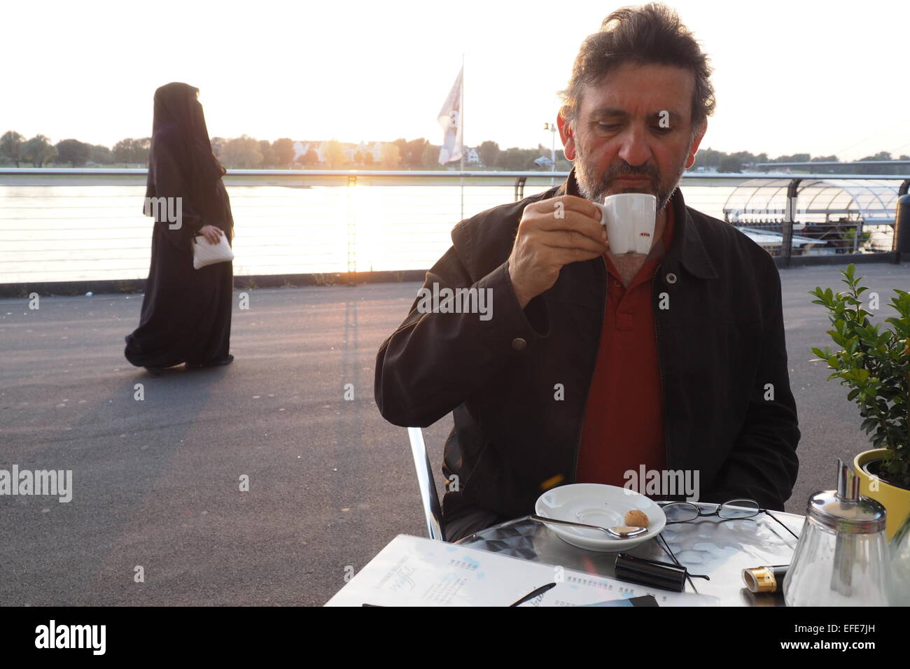 A middle aged man drinking a coffee, with a Muslim woman in traditional dress walking past in the background. Stock Photo
