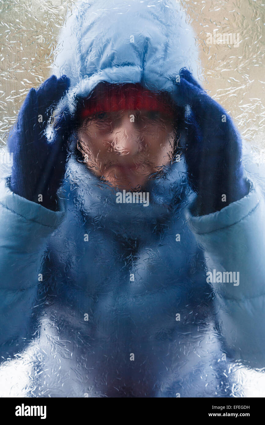 A woman face wearing a warm coat standing outside looking in through behind a frosted glass door wanting to come in from cold. England UK Britain Stock Photo