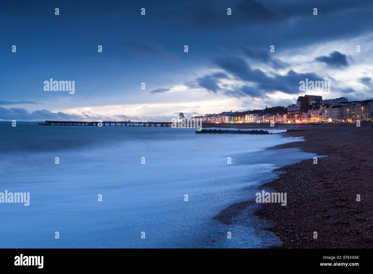 Hastings Seafront and Pier. Waves crash onto the beach at sunset. while the lights of the town twinkle in the background. Stock Photo