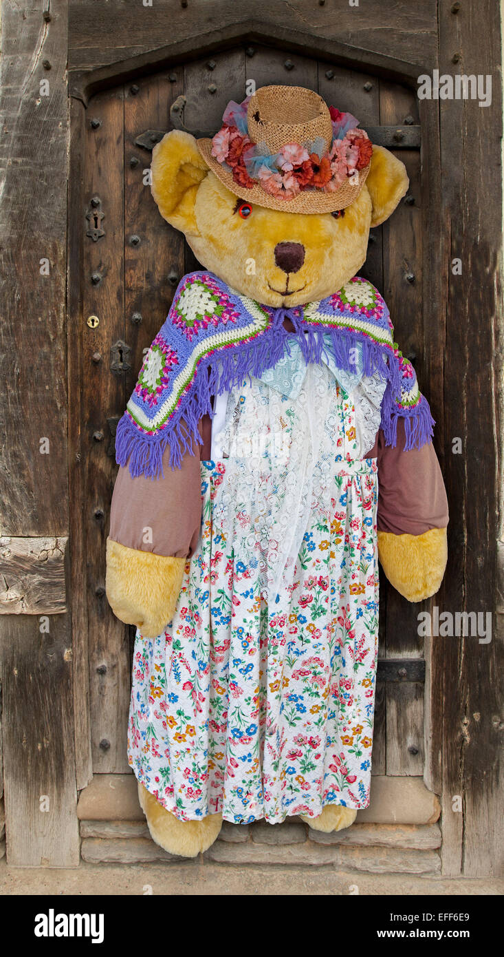 Gigantic life-sized teddy bear wearing floral dress, colourful woollen shawl & straw hat with flowers, against old wooden door of shop Stock Photo