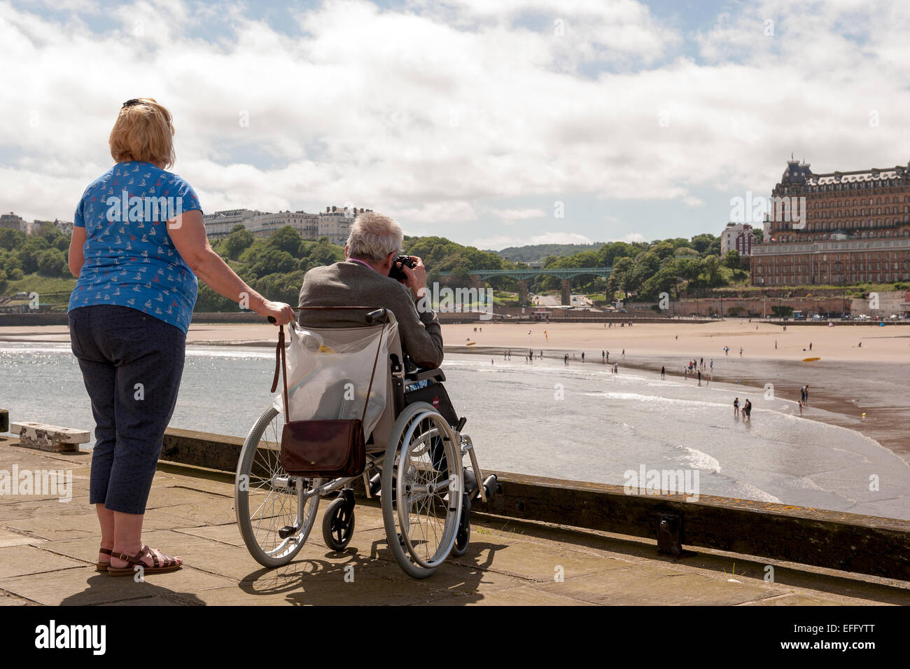 A man in a wheelchair, cared for by a middle aged woman,  takes photographs of the beach and bay at Scarborough, North Yorkshire Stock Photo