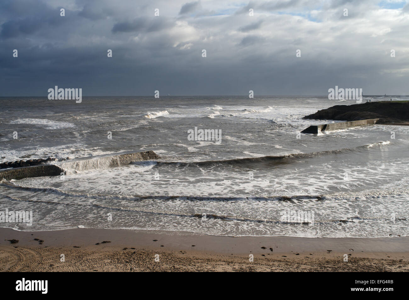 Waves washing over breakwater during stormy conditions, Cullercoats Bay with Tynemouth in background north east England, UK Stock Photo