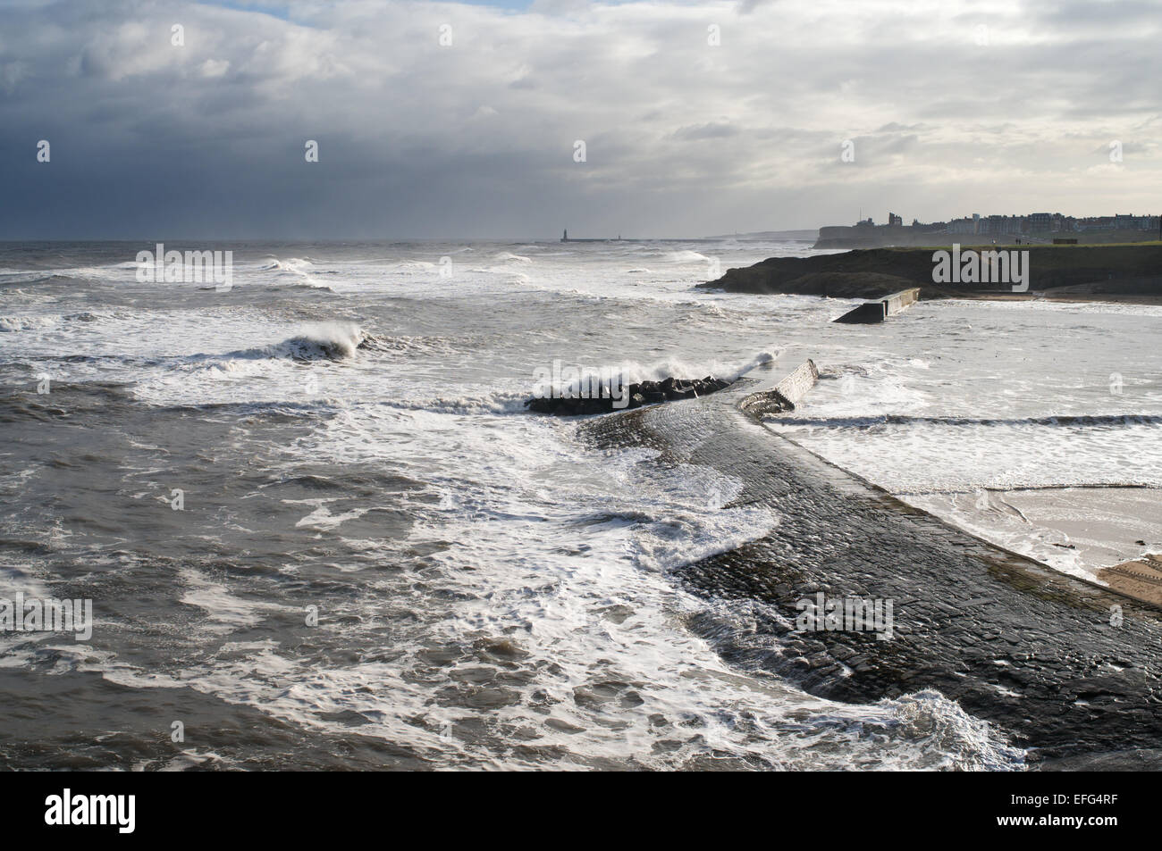 Waves washing over breakwater during stormy conditions, Cullercoats Bay with Tynemouth in background north east England, UK Stock Photo