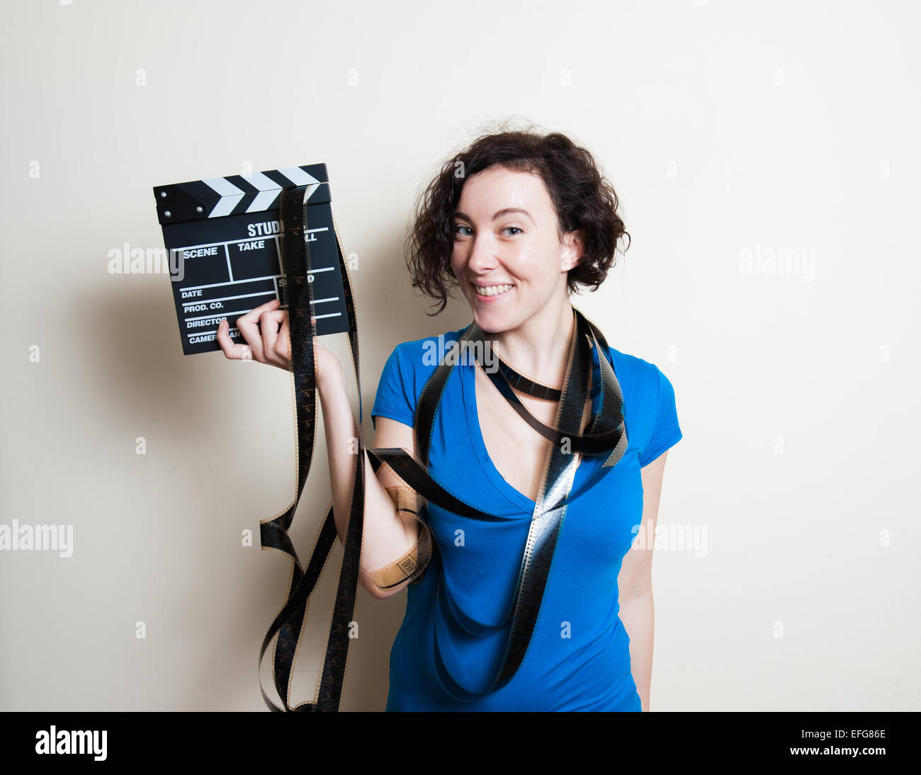 Young pretty woman with blue casual t-shirt and filmstrip around neck is smiling, holding a movie clapper on white background Stock Photo