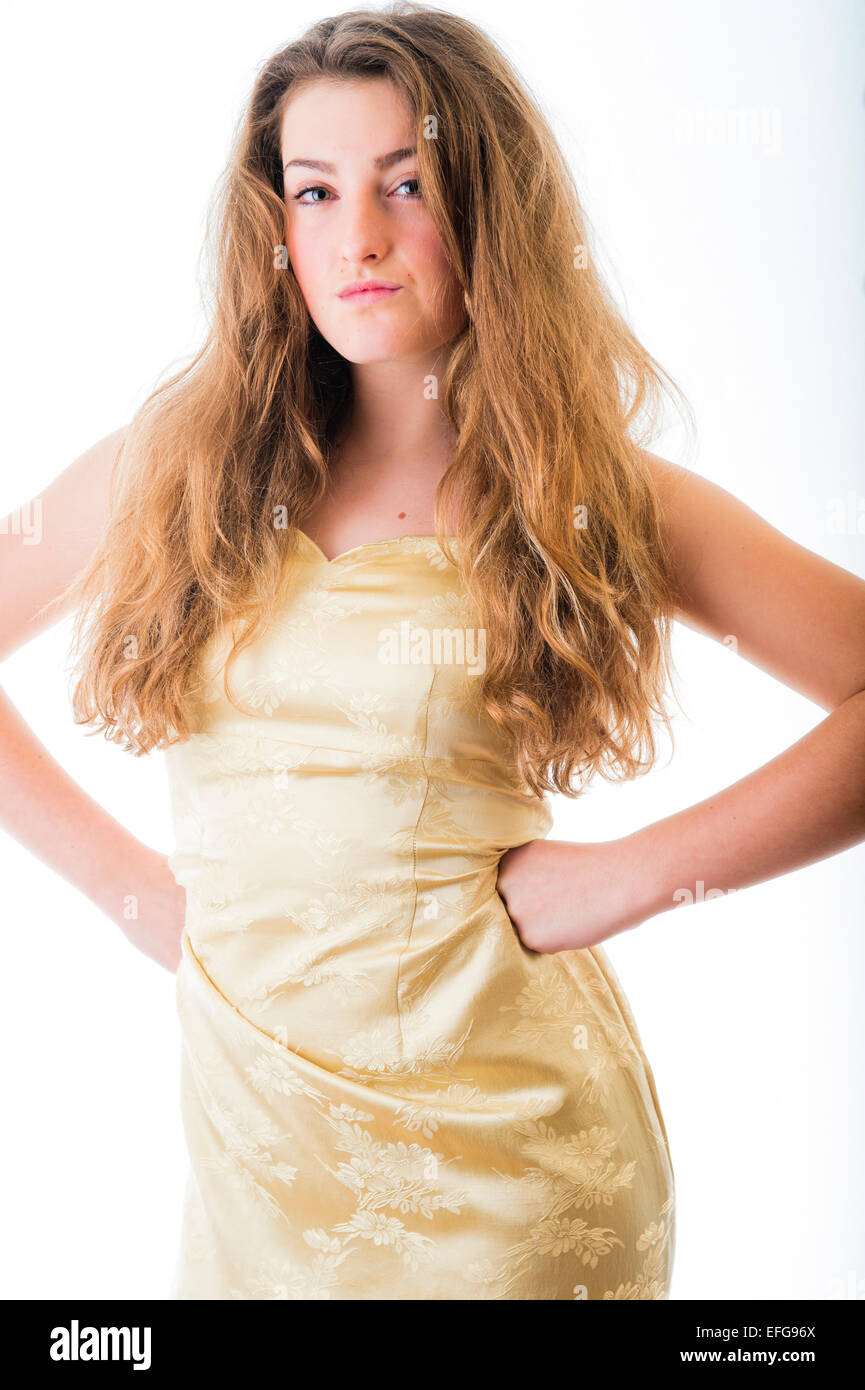 A confident assertive fourteen 14 year old teenage girl with long brown hair, posing in a studio wearing a gold golden dress with her hands on her hips standing facing ahead UK Stock Photo
