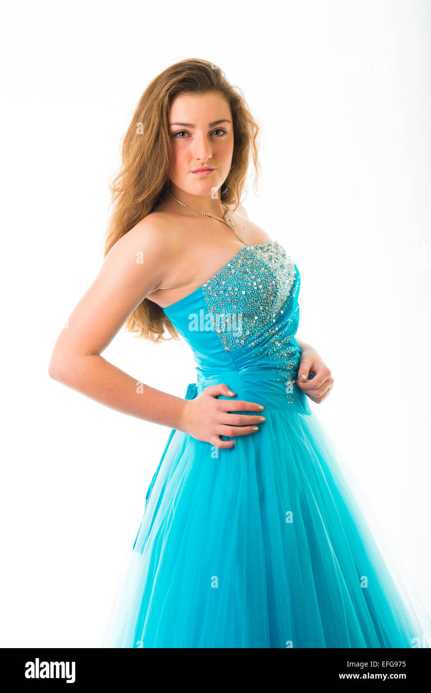 A graceful elegant slim confident fourteen 14 year old teenage girl model with long brown hair, standing with her hands on her hips wearing a blue ball gown party frock dress frock posing in a studio  UK Stock Photo