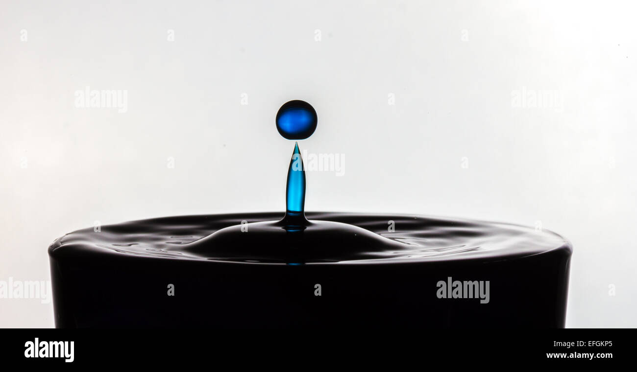 Blue splash of water suspended over glass of black water. The two drops
