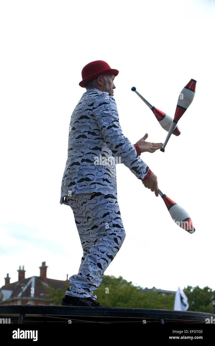 A man in a black and white suit and red hat juggles three clubs in the air. Stock Photo