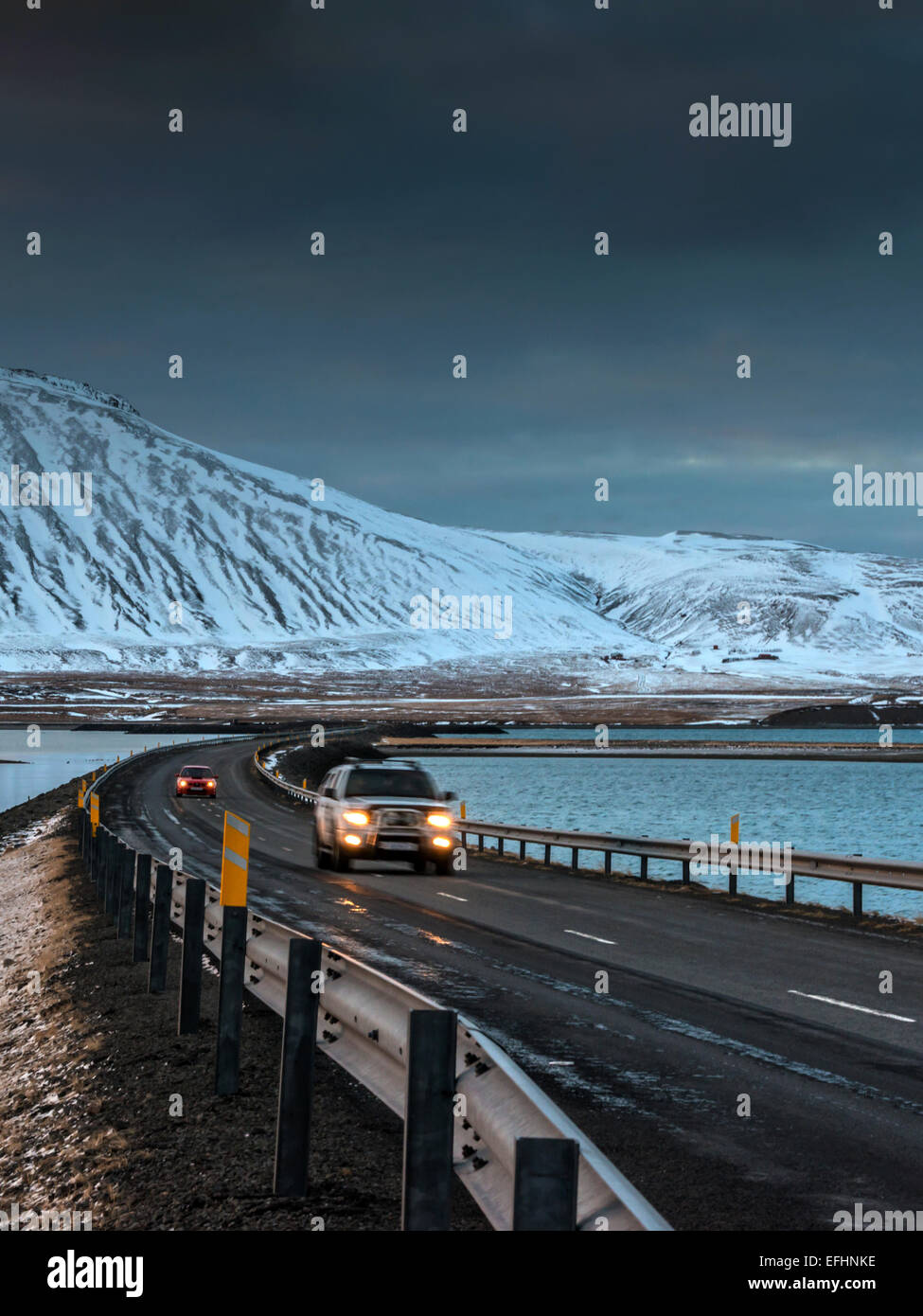 Two vehicles cross the Icelandic causeway at Kolgrafafjorour, with headlights illuminated. Stock Photo