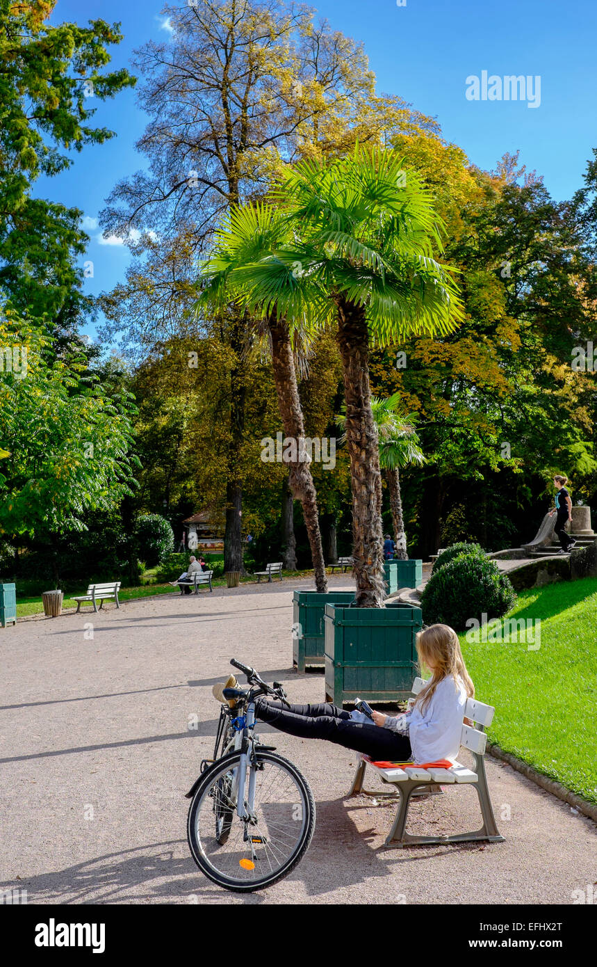 Girl on bench reading a book with legs on her bike Parc de l'Orangerie park Strasbourg Alsace France Europe Stock Photo
