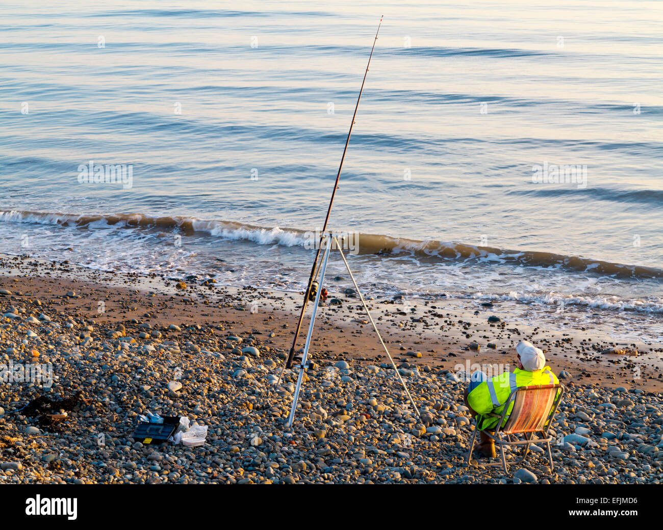 Man sitting in deck chair sea fishing with rod placed on a tripod stand on a beach with pebbles and a calm sea in background Stock Photo