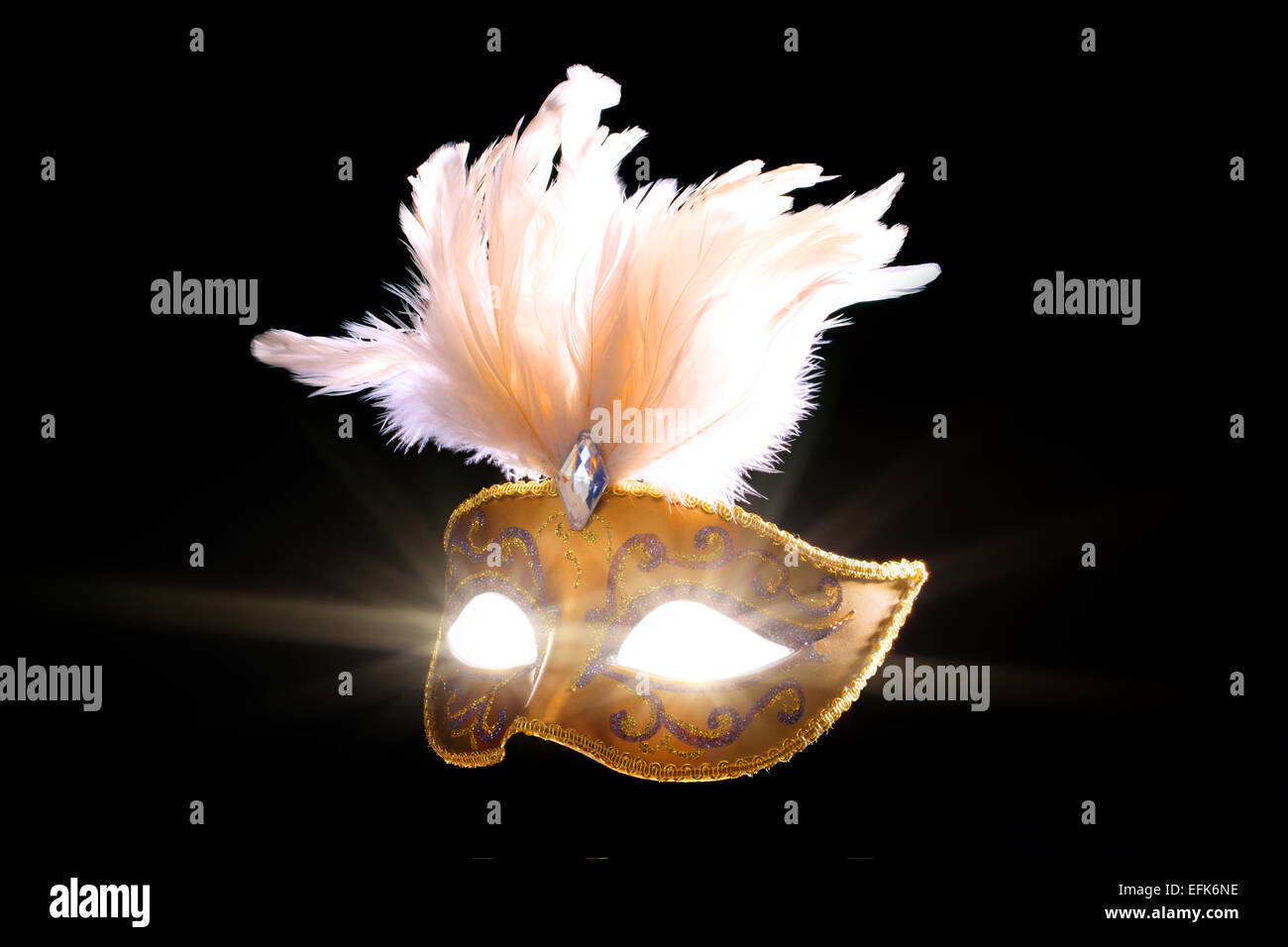 A back lit gold coloured Harlequin mask with flared light emanating from the eyes Stock Photo