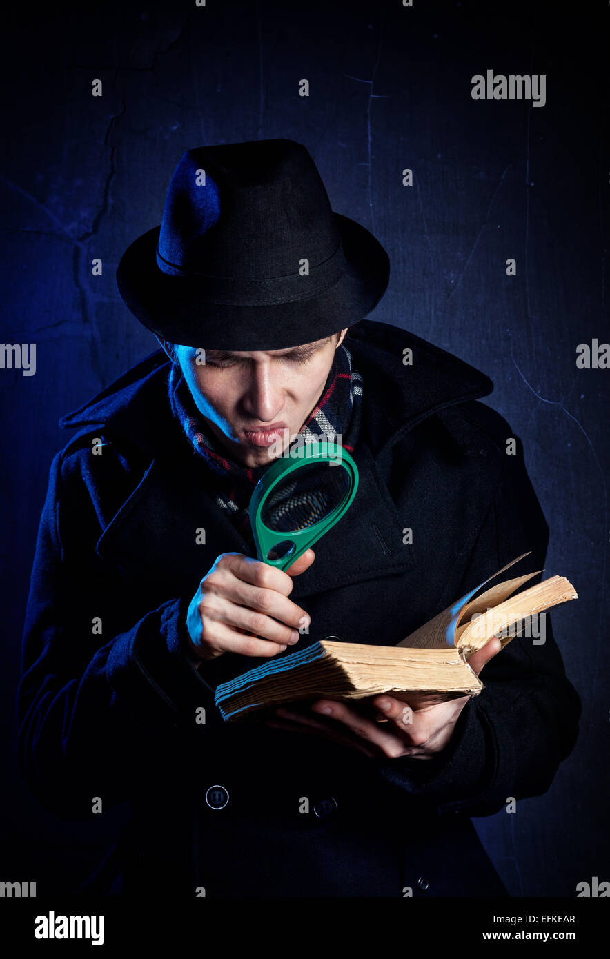 Man in black hat with magnifier glass reading the old book at dark background Stock Photo