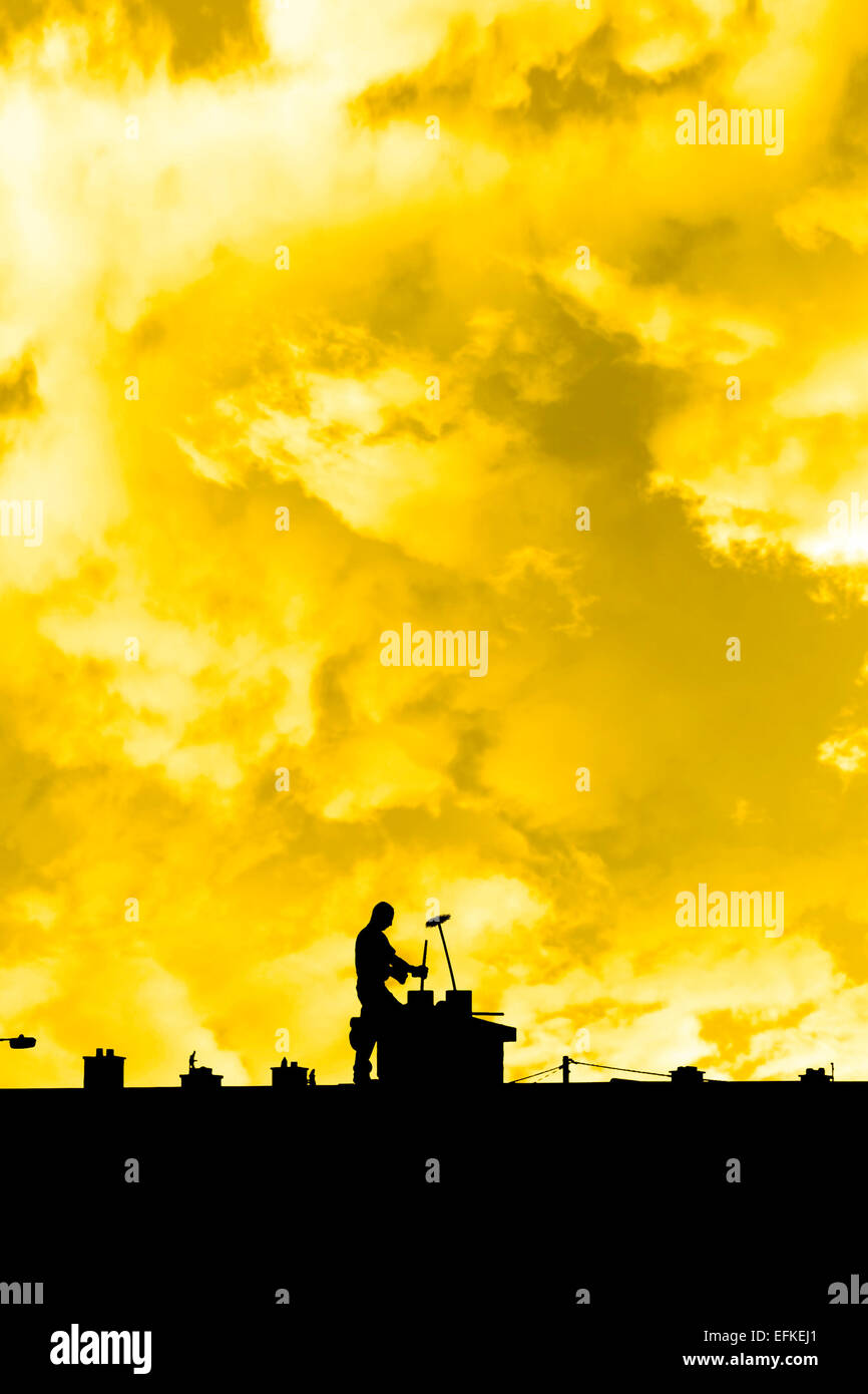 silhouette of a chimney sweep at work on the rooftop of a housing estate with sunset sky in background Stock Photo