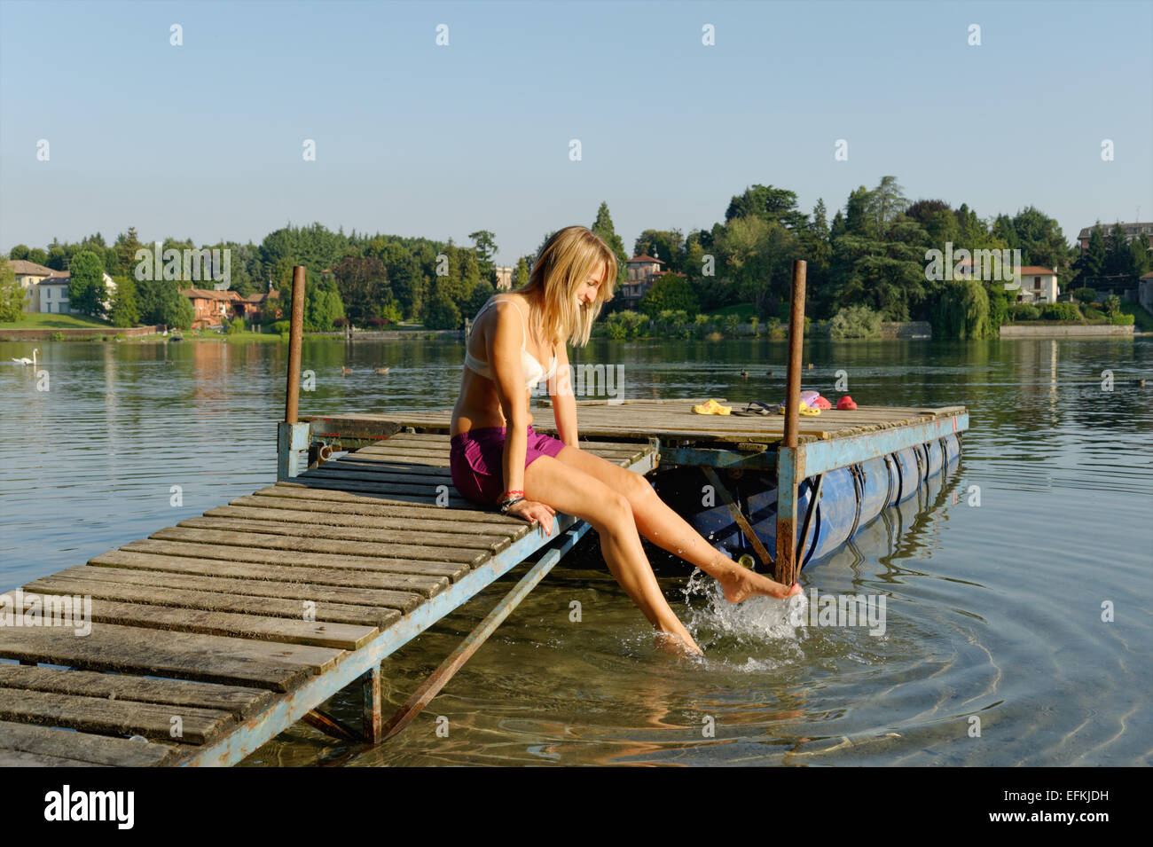 Partially dressed mid adult woman, sitting on jetty, dipping feet in water Stock Photo