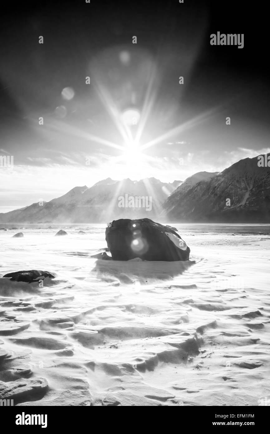 Wind blown snow swirling around a rock on an Alaskan beach with dramatic lens flare in black and white. Stock Photo