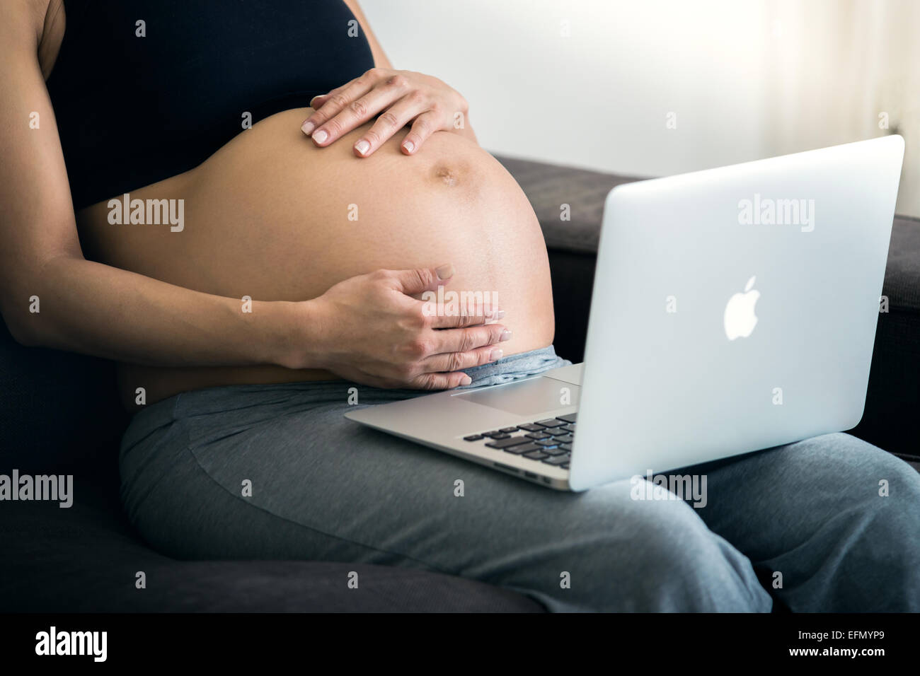 Baby bump, Image of 8 month pregnant woman sitting on a couch, with an imac laptop on her lap, holding her belly. Stock Photo