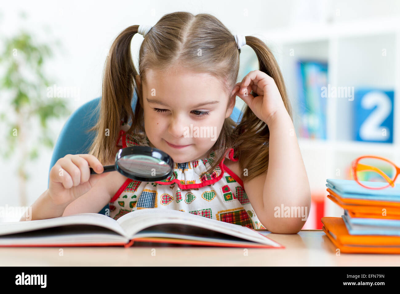 funny child reads book using magnifier sitting at table Stock Photo