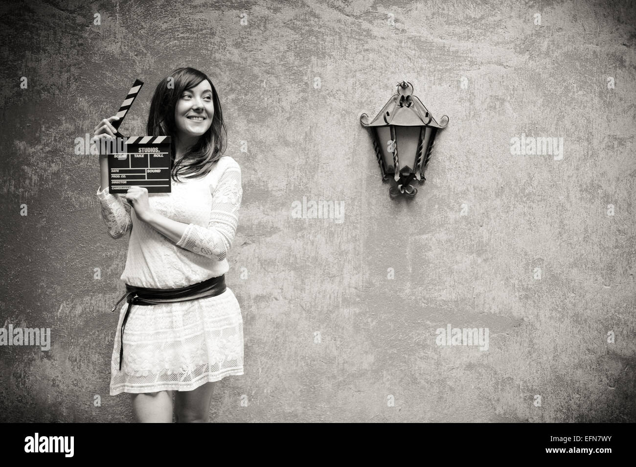 Young woman in 70s hippie style smiling with clapperboard, outdoor wall and lamp background in black and white Stock Photo