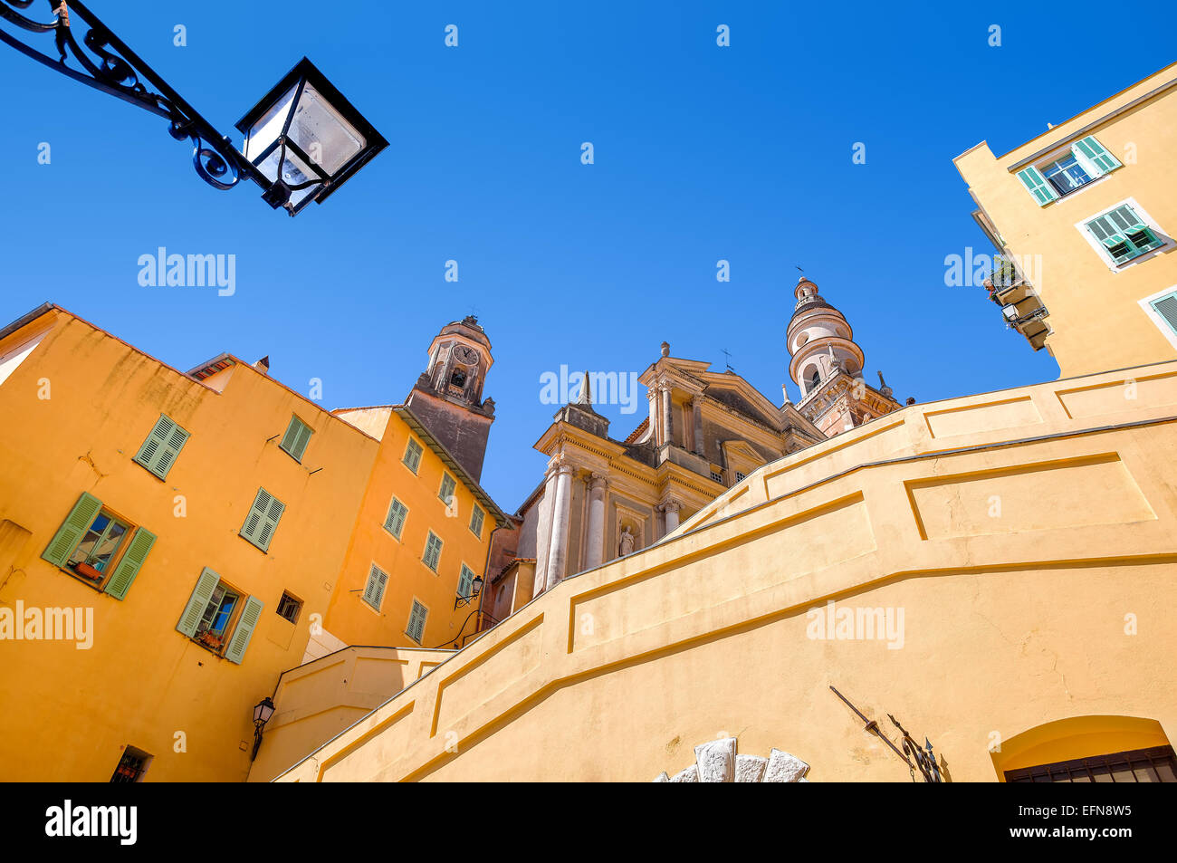 Yellow and orange residential houses and Saint Michel Archange basilica under clear blue sky in Menton, France. Stock Photo