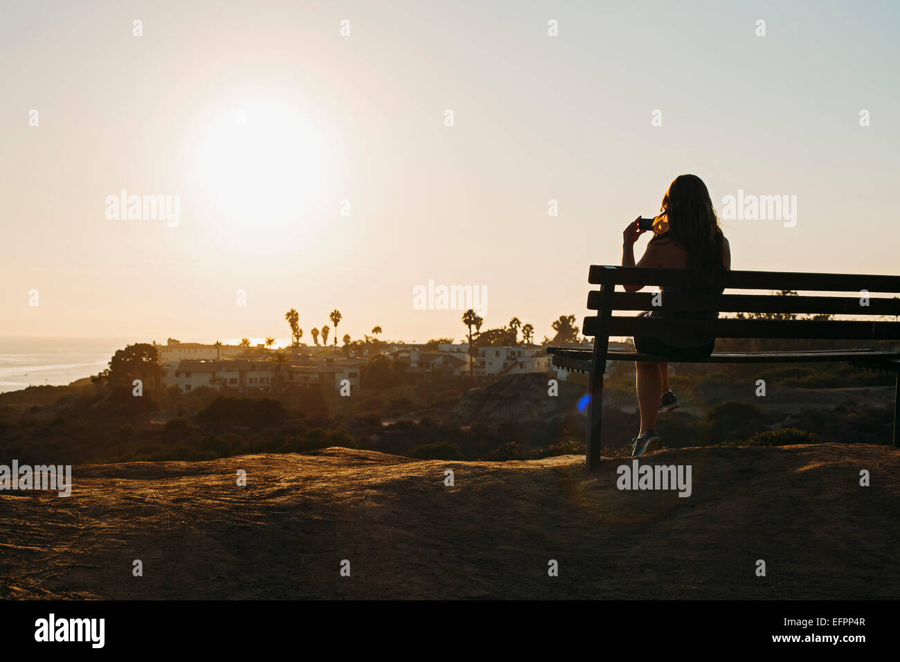Silhouette of young woman sitting on clifftop bench photographing view on smartphone, San Clemente, California, USA Stock Photo