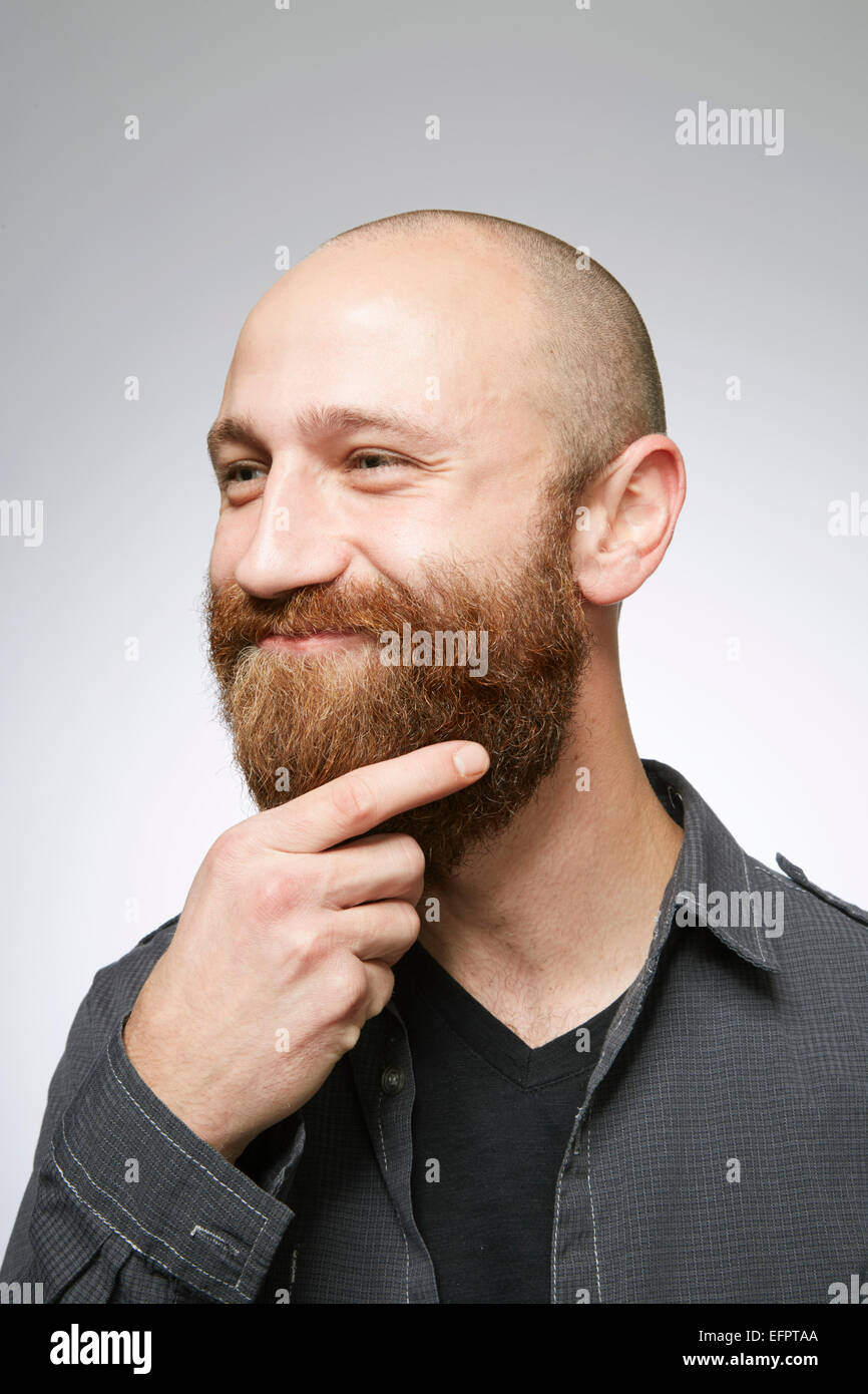 Studio portrait of mid adult man with shaved hair stroking overgrown beard Stock Photo