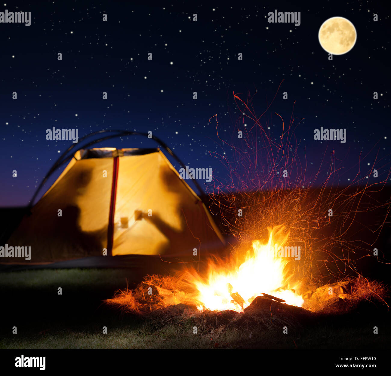Camp shines at night. The campfire in the front as the symbol of adventure and romantic. Stock Photo