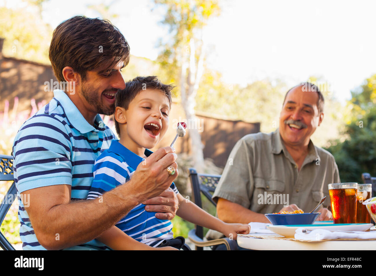 Father feeding son on lap, grandfather in background Stock Photo