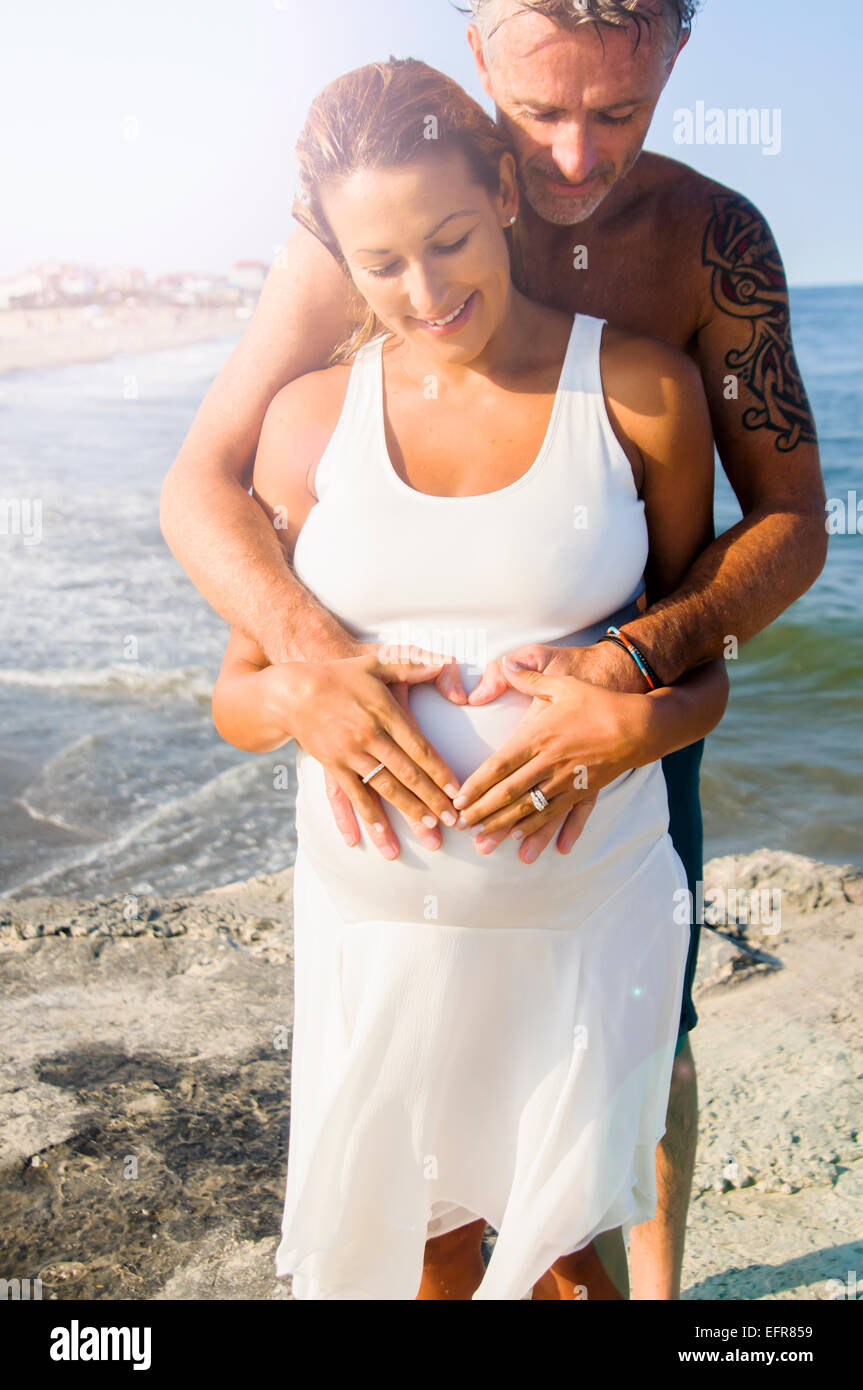 Pregnant mature woman and husband making heart shape on stomach at beach Stock Photo